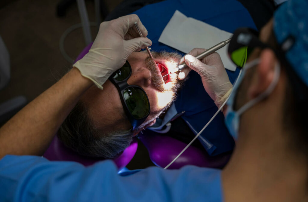 A male dentist is performing a laser treatment on a male patient. The dentist holds laser equipment in his right hand while the patient is lying on a dental chair and wears a protective eyeglasses.