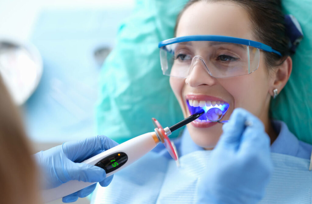 A female patient wearing safety glasses and opening her mouth as a dentist cures one of her teeth with a blue light