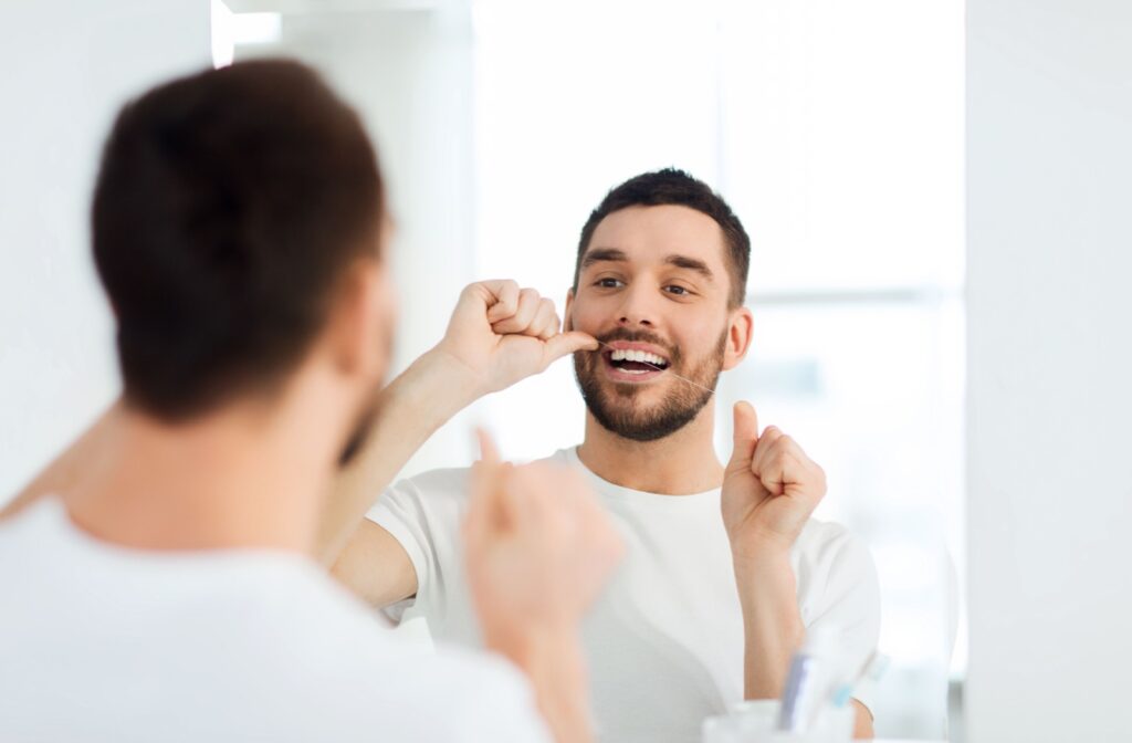 A man flossing his teeth in front of a bathroom mirror.