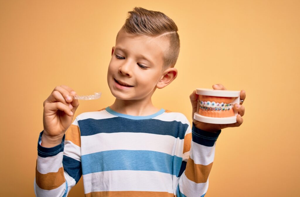 A boy holding and looking at a clear aligner in his right hand while also holding a dental model with braces on the teeth.