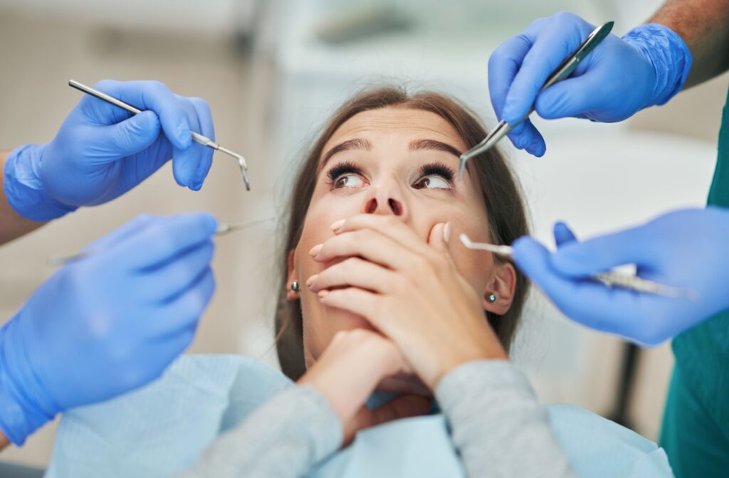 A dental patient covers her mouth with her hand in fear while looking at the instruments held by her dentist and hygienist.