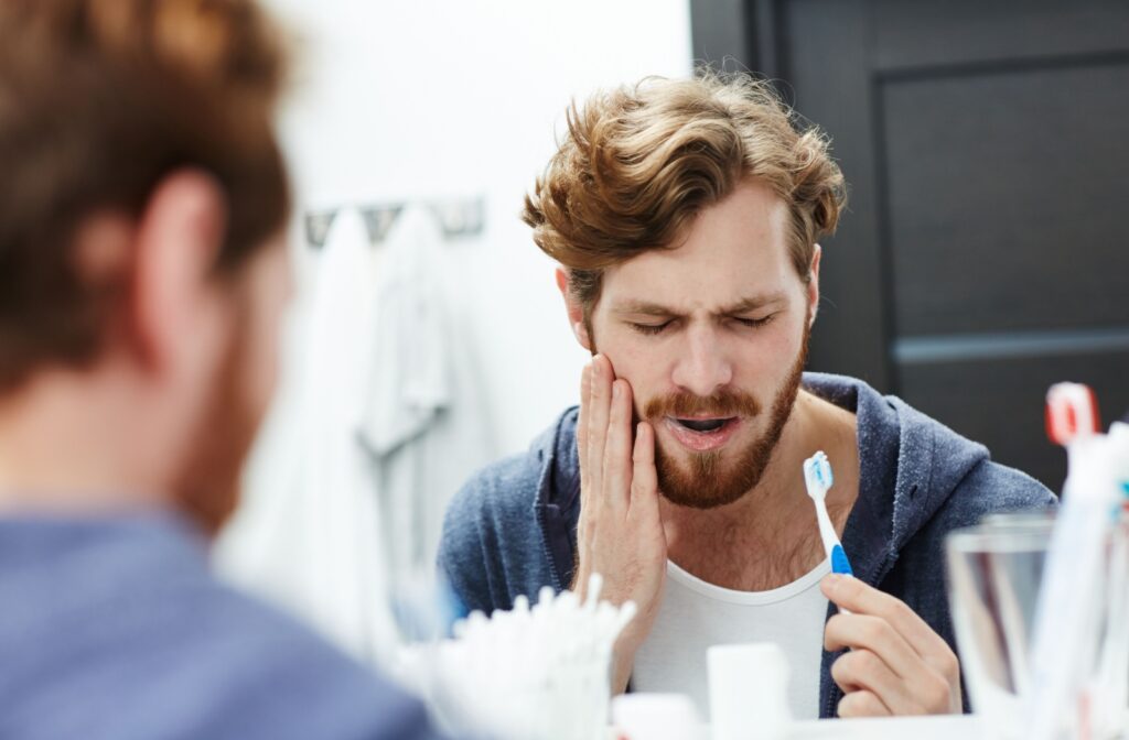 A young adult wincing in the mirror while brushing their teeth due to sudden pain from brushing their teeth too much.