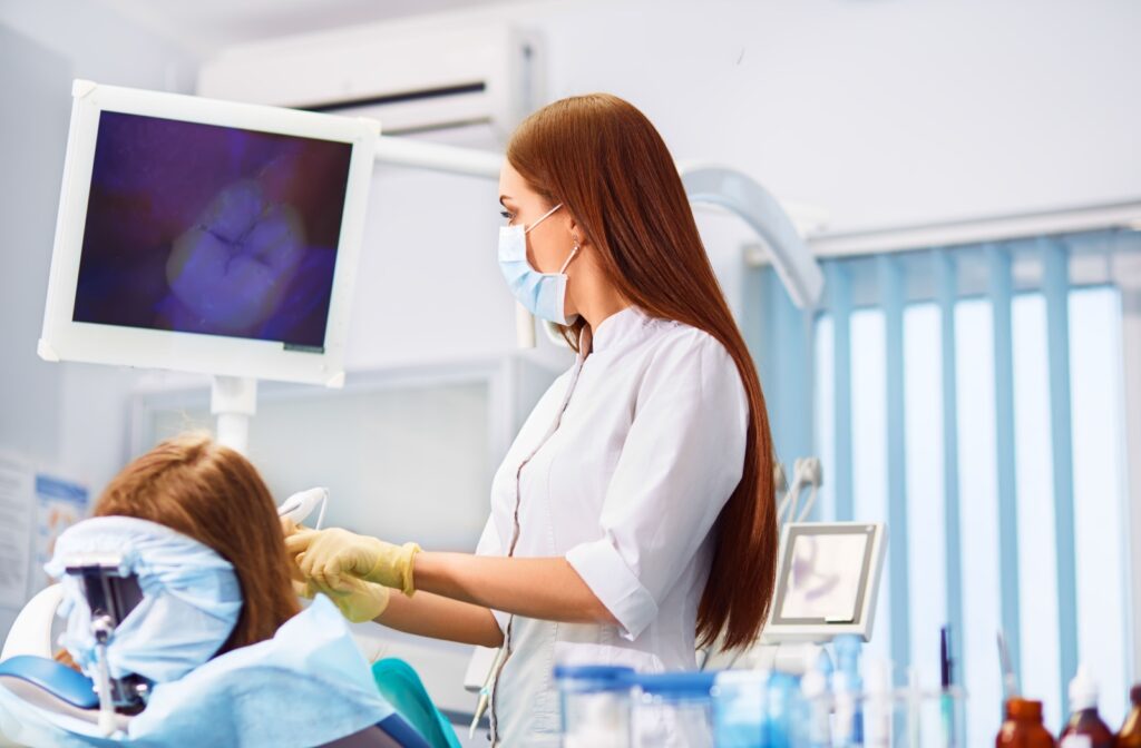 A dentist wearing a mask and gloves looks at a screen displaying a tooth image while examining a patient in the dental chair.