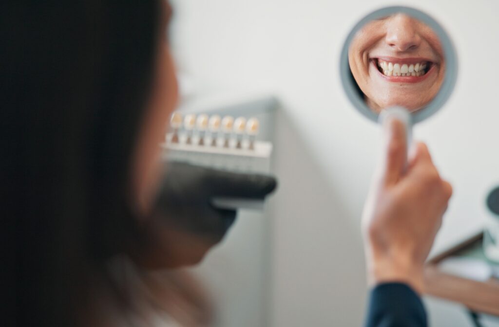 A person looking at their teeth in a mirror and smiling as a dentist hold dental crowns in front of them