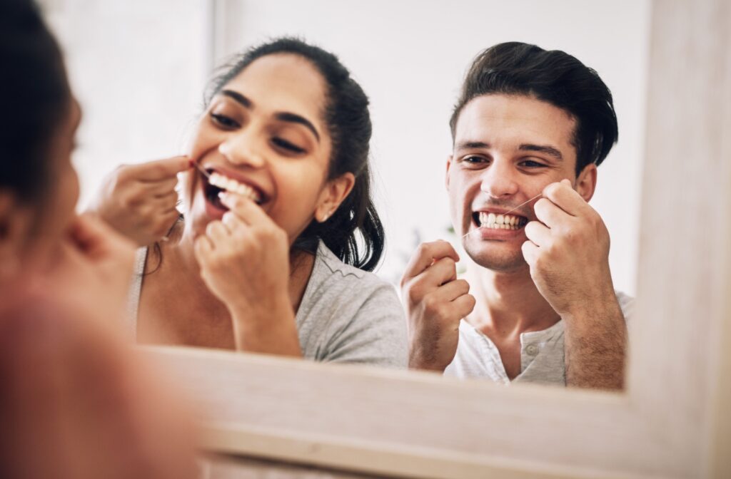 Two people flossing their teeth together as they look into a shared mirror